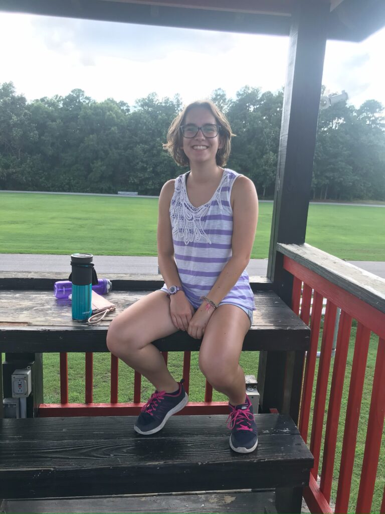 Girl with shoulder length hair sitting on a wooden structure overlooking a field