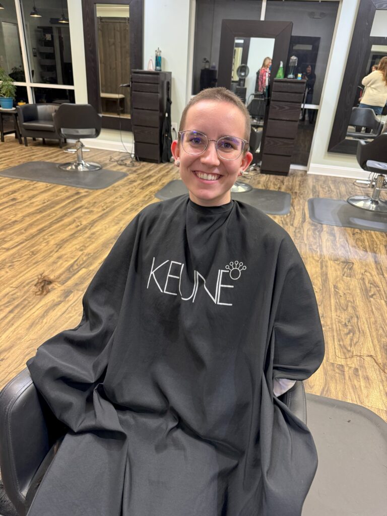 A woman sitting in a salon chair, smiling with a shaved head.