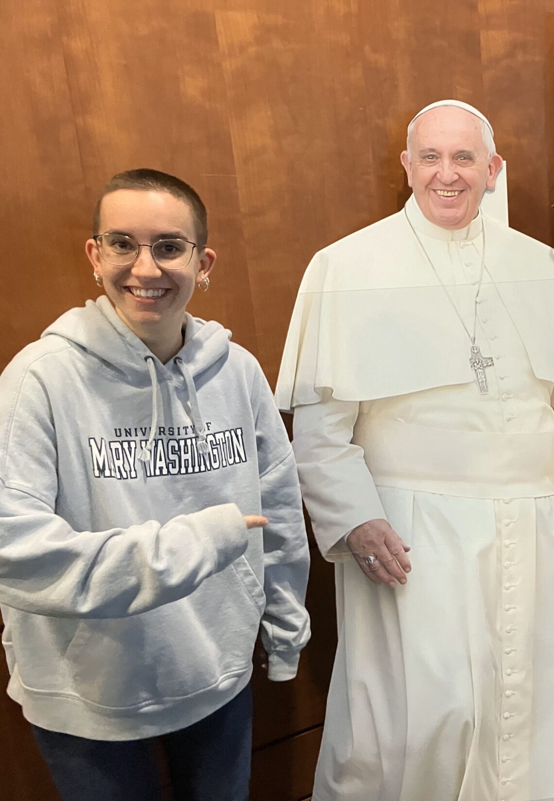A smiling woman standing next to and pointing to a cardboard cutout of Pope Francis.