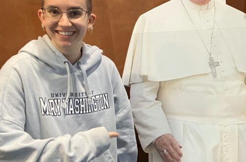 A smiling woman standing next to and pointing to a cardboard cutout of Pope Francis.