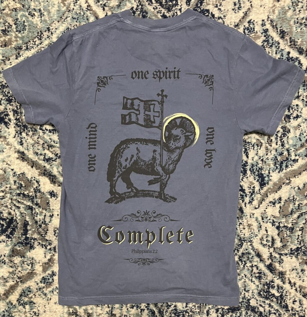 The back of a blue t shirt with a box design with the words "one mind" on the left side, the words "one spirit" on the top, the words "one love" on the right, and the word "complete" on the bottom with the reference for the Bible quote underneath, which reads "Philippians chapter 2 verse 2." There is a drawing of a lamb in the middle of the box, with a halo around its head and it is carrying a flag with a cross on it.