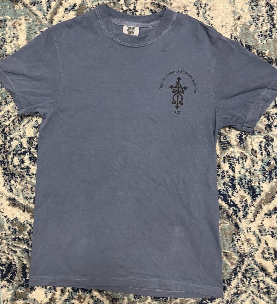 The front of a blue t shirt bearing the words "Catholic Campus Ministry Summit 2025." There is a logo of a cross as well as the Greek symbols for alpha and omega