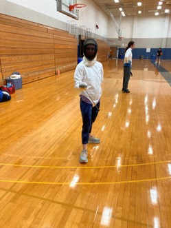 A woman in fencing gear pointing a sabre at the camera.