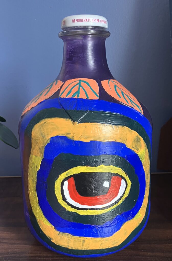 Acrylic paint on an apple cider jar. From the front view there is a red eye with concentric circles expanding out from the eye. The circles are alternating in dark green, dark blue, and yellow.