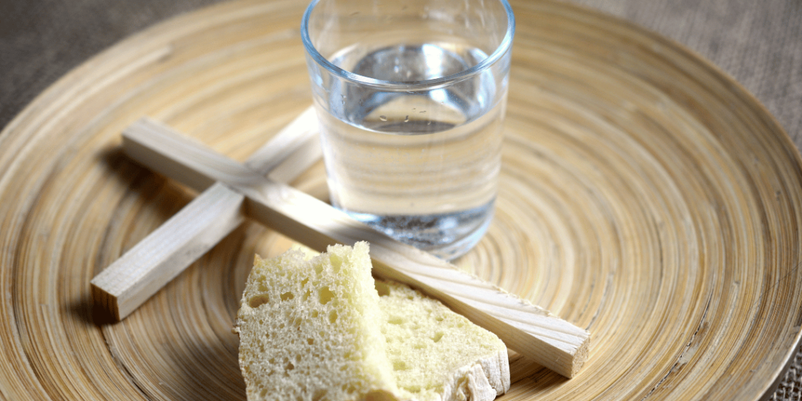 A wooden plate with a cup of water, a piece of bread, and a wooden cross on it.