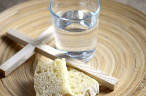 A wooden plate with a cup of water, a piece of bread, and a wooden cross on it.