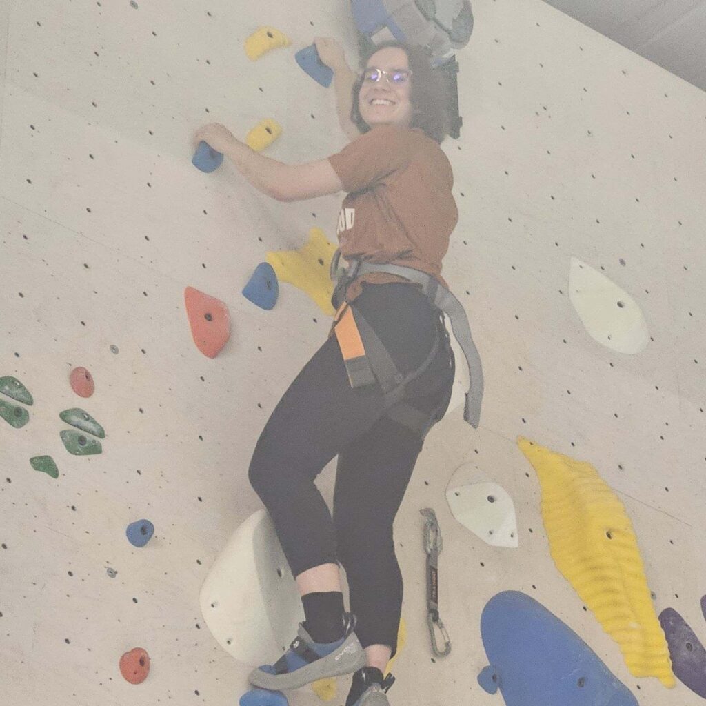 A young woman smiling at the top of an indoor rock climbing wall.