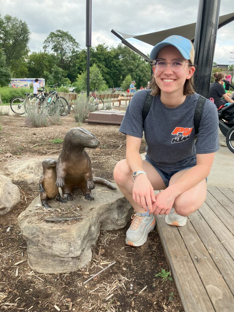 A young woman squatting beside a statue of a mother otter and her pup.