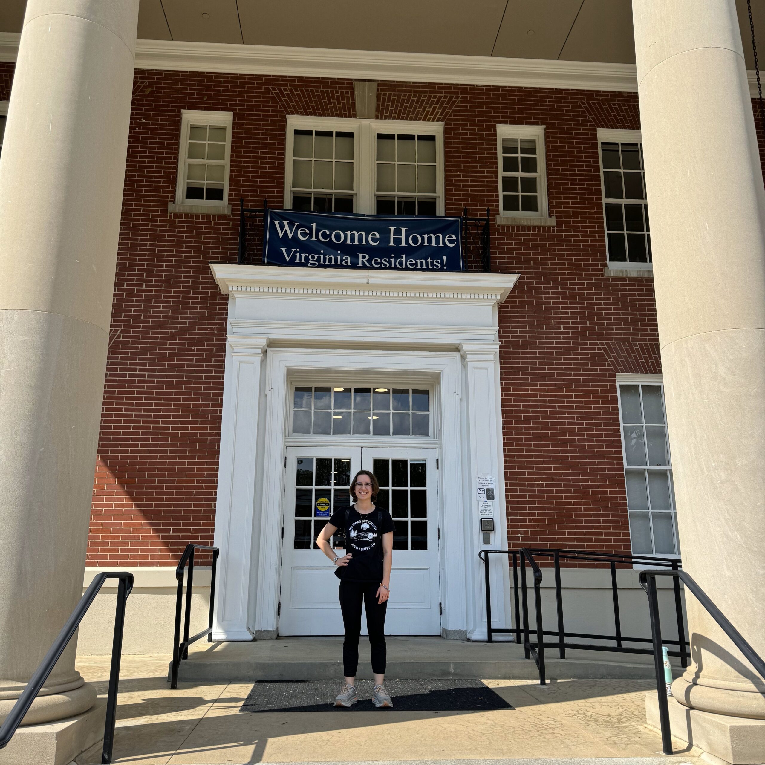 A young woman standing in front of a building with a banner above the front doors that reads, "Welcome Home Virginia Residents!"