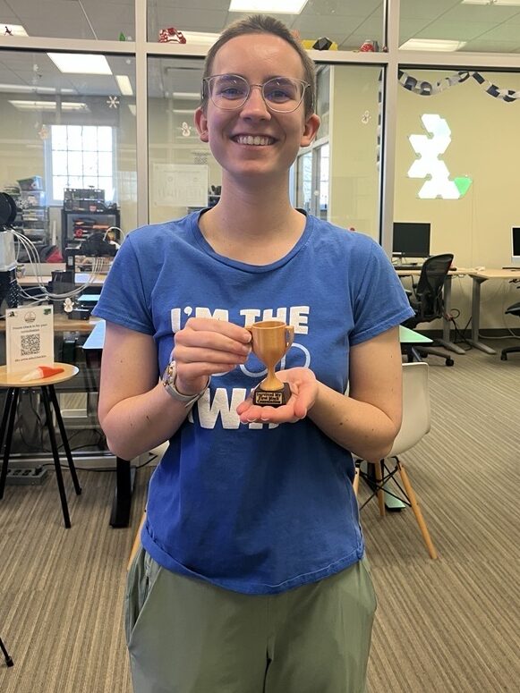 A young woman smiling while holding a 3D-printed trophy that says, "I survived my first mock consultation."