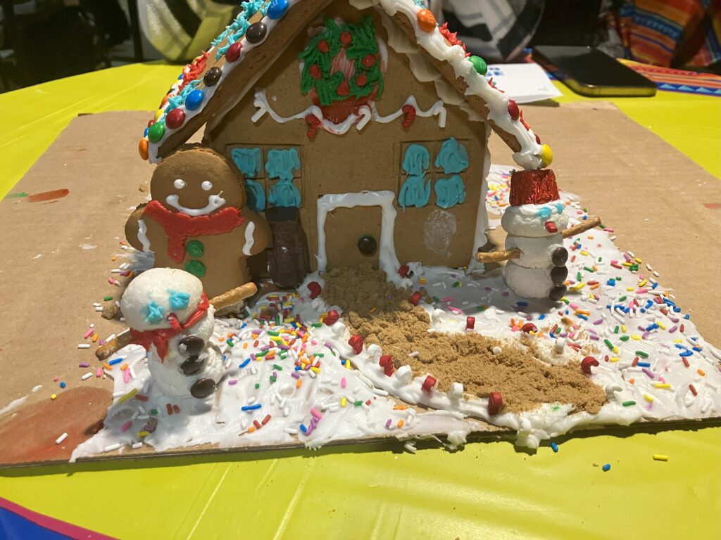 A fully decorated gingerbread house with two snowmen in front of the house made out of marshmallows.